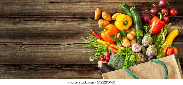 Beige canvas grocery bag with dark green handle fallen over while dropping vegetables and fruits on wood plank
