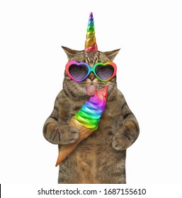 The beige cat unicorn in heart shaped sunglasses is licking a rainbow ice cream cone. White background. Isolated.