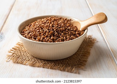 Beige ceramic bowl full of raw wholegrain buckwheat groats and spoon on a white wooden table. Cooking buckwheat porridge. Concepts of gluten free diet and vegetarian food. Close-up.