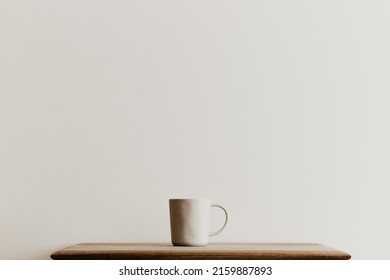 A Beige Ceramic Mug On A Brown Wooden Board In Front Of A White Wall. Hand Made Coffee Cup With Copy Space. Minimal Concept, Simple Design. 