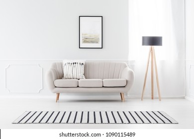 Beige couch standing against white wall with molding and watercolor poster in cozy living room interior