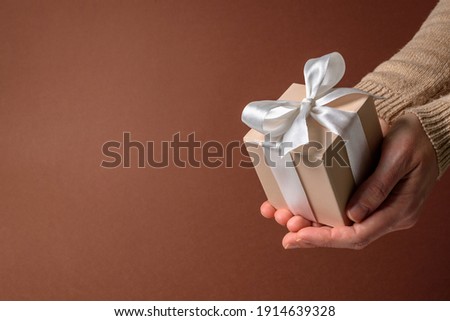 Beige gift box with white bow in woman hands on brown background