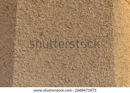 Beige plaster wall with rough texture illuminated by natural sunlight. Architectural detail showing decorative facade surface with sand and cement mix.