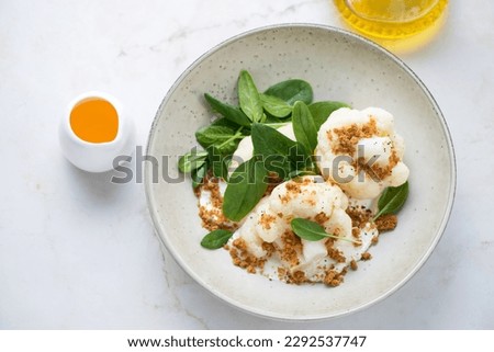 Beige plate with steamed cauliflower florets, ricotta and fresh spinach, high angle view on a light-beige marble background, horizontal shot