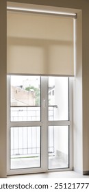 Beige roller blind is pulled down to cover a large glass door that leads to a balcony. The door is framed with white trim and the wall is a pale beige color.