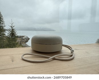 Beige round Bluetooth speaker with a strap on a wooden shelf by a panoramic window, showing a serene view of trees, mountains, and a fjord, blending modern design with natural beauty.