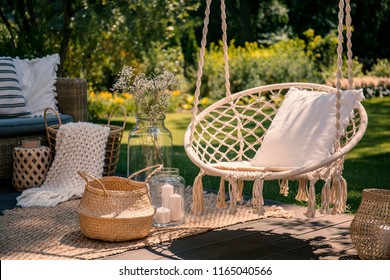 A Beige String Swing With A Pillow On A Patio. Wicker Baskets, A Rug And A Blanket On A Wooden Deck In The Garden.