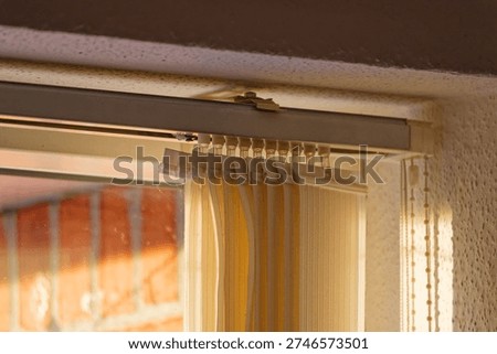 Beige vertical blinds installed on a window frame with sunlight casting shadows and a blurred brick wall visible outside the window.