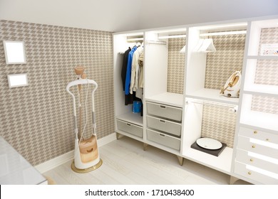 Beige Wardrobe For Clothes In The Dressing Room. Open Shelves, Things Hang On A Hanger. On The Shelves, Scales, Iron, Hangers. On The Floor Is A Garment Steamer. Picture Frames.