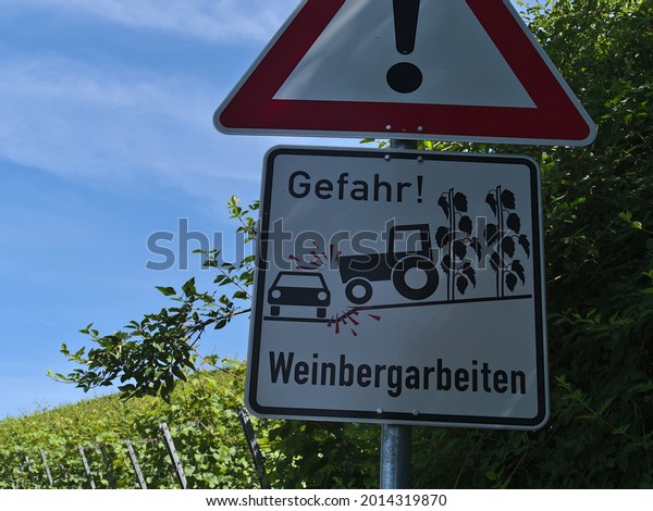 Beilstein, Germany - 07-03-2021: Metal road\
sign with red frame and exclamation mark warning of accidents that\
could be caused by works in the vineyards with wine plants in\
background.