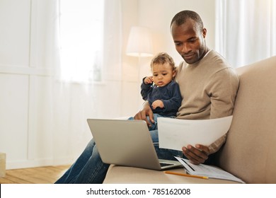 Being a daddy. Concentrated dark-eyed bearded afro-american man holding his son on his lap while working on the laptop and holding a sheet of paper