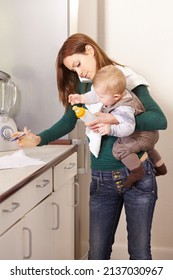 Being A Mom Is A Real Juggling Act. Young Mom Trying To Multi-task While Holding Her Baby Son And His Bottle.