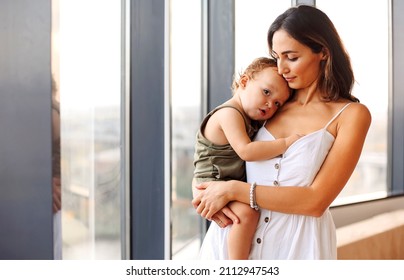 Being mom. Young beautiful woman mother holding cute baby boy while standing by window at home. Woman in white dress cuddling her little son infant and expressing love and care - Powered by Shutterstock