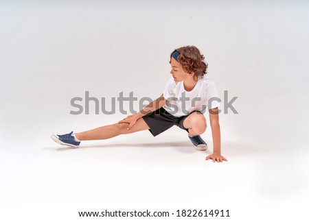 Being physically active. Full length of caucasian teenage boy in sportswear stretching legs, warming up before training isolated over grey background in studio
