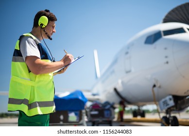 Being responsible at work. Side view of aviation technician noting data before the flight. Blue sky and passenger plane on blurred background