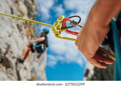 Belay device close-up shot with a boy on the cliff climbing wall. He hanging on a rope in a climbing harness and his partner belaying him on the ground. Active people and sports concept image - Powered by Shutterstock