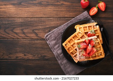 Belgium waffles. Homemade waffles with strawberries, powdered sugar and cup of coffee on black plate on old wooden table background. Breakfast. Top view. Mockup for design idea.