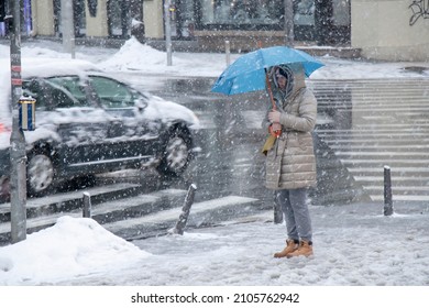 Belgrade, Serbia - January 11, 2022: Woman Waiting At Crossroad Under Blue Umbrella On A Snowy City Street During Heavy Snow Fall