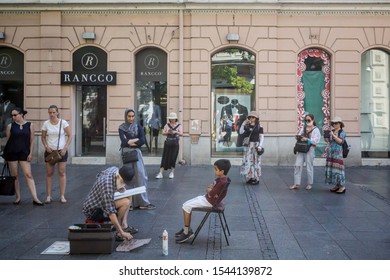 BELGRADE, SERBIA - JULY 6, 2019: Street Cartoonist Drawing The Caricature Of A Children In Kneza Mihailova, The Main Street Of Belgrade, Serbia, Surrounded By Tourists Observing Him

