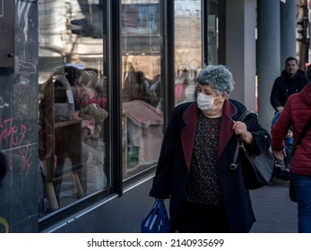 BELGRADE, SERBIA - MARCH 19, 2022: Selective Blur On An  Old Woman Looking At Prices In Window Of Shop On A Street Of Belgrade Wearing Facemask During Coronavirus Covid 19 Crisis.

