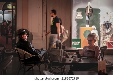 BELGRADE, SERBIA - SEPTEMBER 5, 2021: Selective Blur On A Senior Man Sitting At Night Drinking Beer Outdoor At A Bar Cafe, With A Young Man Rushing Behind With A Speed Blur, In Belgrade.

