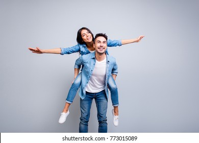 I believe I can fly. Love story of attractive, funny, cheerful couple - handsome man carrying his lover on back like plane, woman opens her hands to the side over grey background