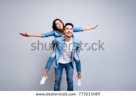 I believe I can fly. Love story of attractive, funny, cheerful couple - handsome man carrying his lover on back like plane, woman opens her hands to the side over grey background