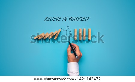 Believe in yourself sign over a hand drawn man stopping falling dominos in a conceptual image. Over blue background.