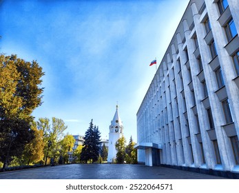 A bell tower stands beside a modern building, framed by colorful trees on a sunny day. - Powered by Shutterstock