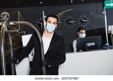 Bellboy In Medical Mask Pushing Luggage Cart Near Blurred Administrator In Hotel Lobby