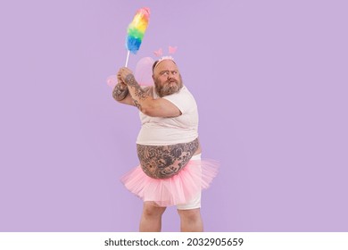 Bellicose Mature Actor Man With Overweight In Carnival Fairy Suit With Pink Wings Holds Dust Brush On Purple Background In Studio