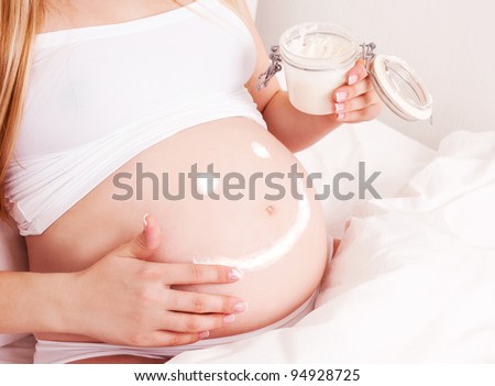 belly of a  young pregnant woman sitting in bed at applying cream  at home