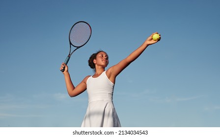 From below African American female athlete in white dress preparing to hit ball with racket against blue sky while playing tennis in daytime - Powered by Shutterstock