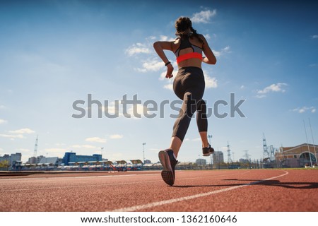 From below back view of athletic teen girl in sportswear with fitness tracker on her wrist running on track at stadium on summer day