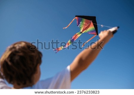 From below back view of unrecognizable kid with multicolored rainbow kite flying against cloudless blue sky in sunny summer day