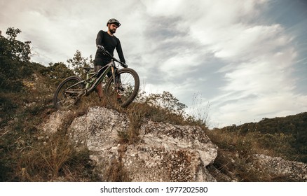 From Below Bearded Man In Helmet Pushing Bicycle And Walking Down Rocky Hill Against Cloudy Sky In Nature