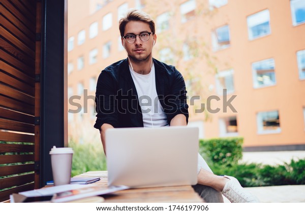 From below of calm male\
in casual wear and glasses with earphones using laptop working on\
freelance project listening to music sitting on bench in street\
looking at camera