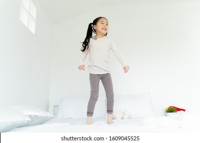 From Below Of Cheerful Adorable Asian Girl In Casual Wear Playing In Bedroom Glad Smiling Kid Jumping On Bed In House