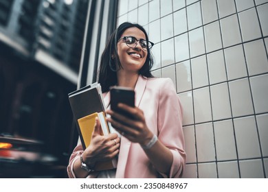 From below of cheerful busy woman in eyeglasses and blazer using mobile phone and smiling excited with great news while standing next to wall of building with paper folder and diary in hand