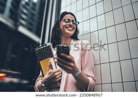 From below of cheerful busy woman in eyeglasses and blazer using mobile phone and smiling excited with great news while standing next to wall of building with paper folder and diary in hand
