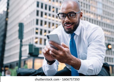 From Below Cheerful Elegant Bald African American Executive Man In White Shirt And Blue Tie Browsing Mobile While Sitting On Street Stairs On Background Of Blurred Business Center 