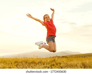 From Below Of Cheerful Little Child Girl In Casual Clothes Having Fun And Jumping With Raised Arms In   Field In Summer Evening
