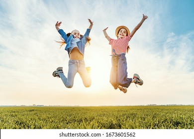 From Below Of Cheerful Little Sisters In Casual Clothes Having Fun And Jumping With Raised Arms In Green Field In Summer Evening
