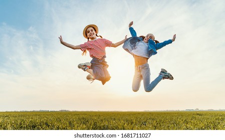 From Below Of Cheerful Little Sisters In Casual Clothes Having Fun And Jumping With Raised Arms In Green Field In Summer Evening
