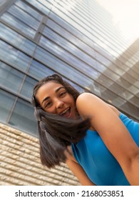 From Below Cheerful Young Hispanic Brunette Bending Forward And Looking At Camera With Smile Against Contemporary Building With Glass Walls On City Street