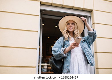 From below curly blonde woman in light dress denim jacket and straightening hat holding mobile phone in hand leaving cafe - Powered by Shutterstock