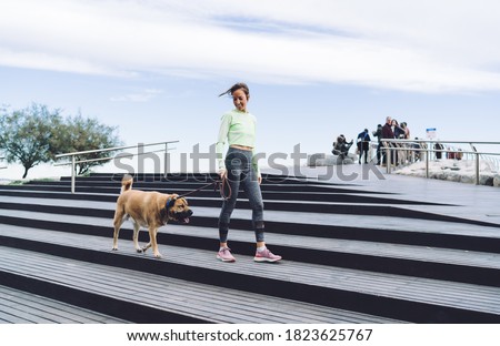 From below of full body cheerful female in activewear walking dog on leash down street steps of city located near sea
