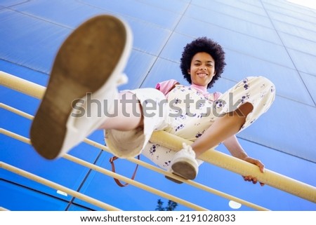 From below full body of positive African American female in overall looking at camera while sitting on fence near glass building