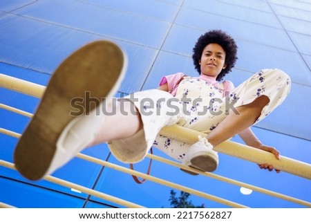 From below full body of young African American female in casual outfit looking at camera with serious face while sitting on railing