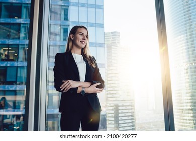 From below of glad businesswoman in formal wear standing with folded arms near window while having break and looking away - Shutterstock ID 2100420277